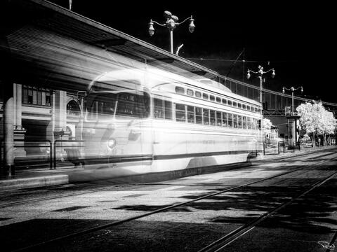 A black-and-white, motion-blurred historic San Francisco streetcar departs the station.